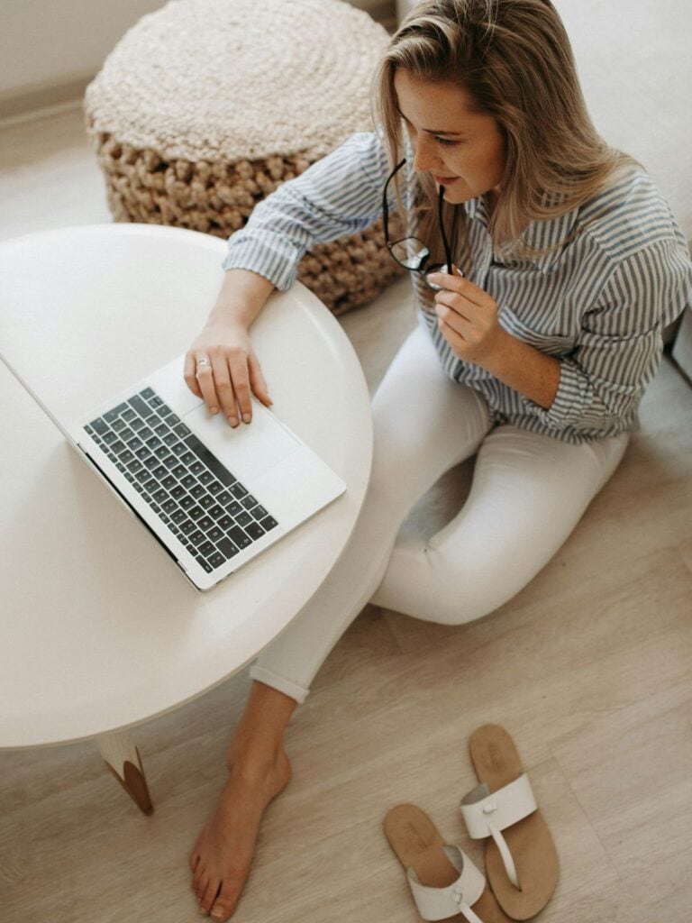 A woman sitting on the floor holding a pair of glasses and working on a computer.