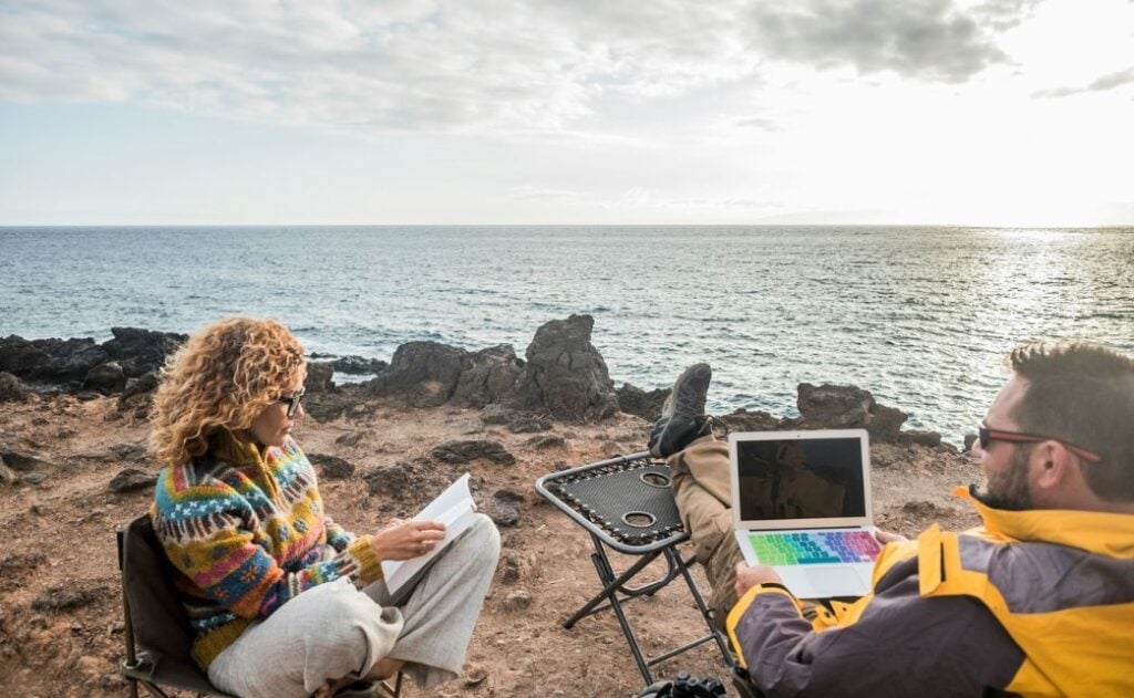 A man and a women sit on chairs at the beach. The man is holding a laptop and the woman is reading a book. Bloggers actually make money, even from the beach.