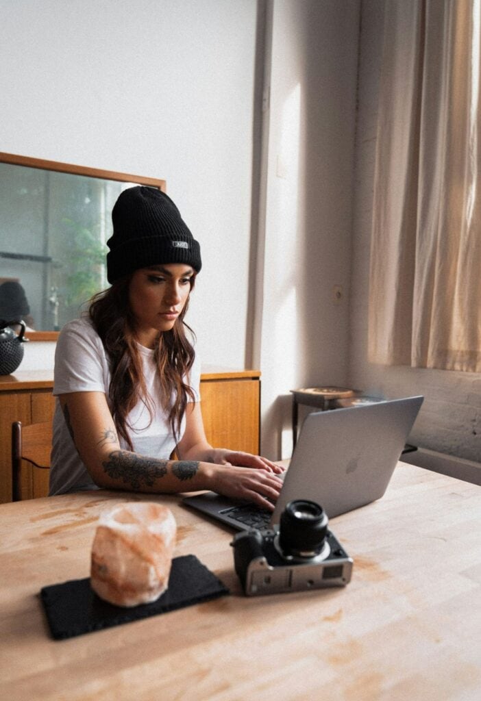 A woman in a black hat sits at a computer typing next to a camera. 