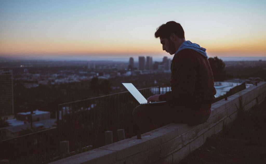 A man sitting at dusk with a laptop on his lap and a city skyline behind him in the background. 