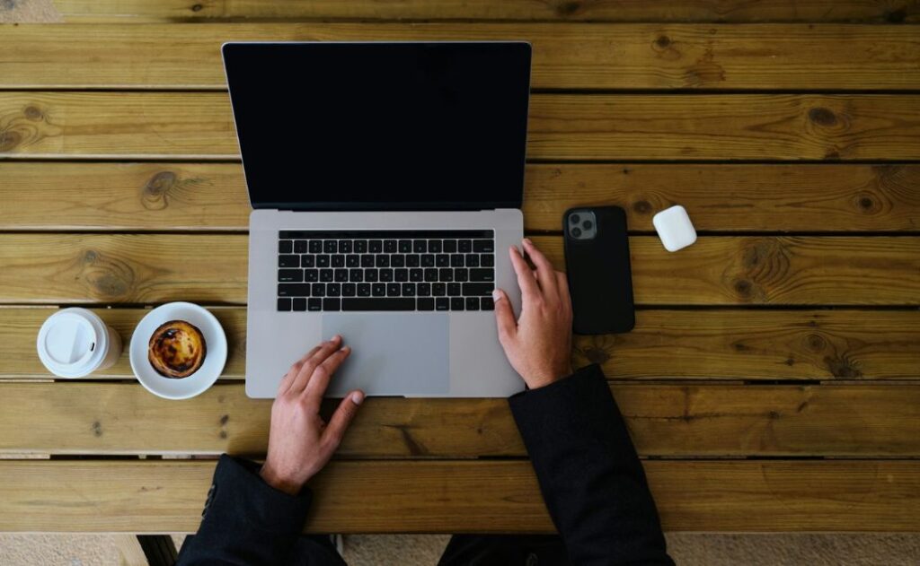 two hands at a laptop with a smartphone and a muffin next to it. 