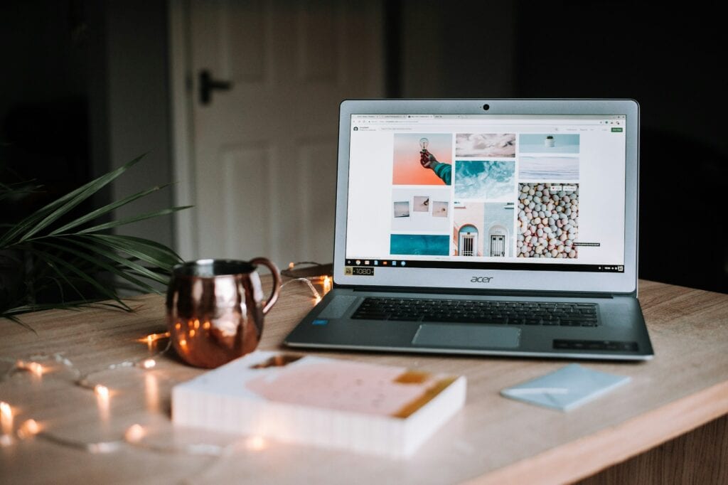 A laptop open on a desk next to a book and a copper cup. 