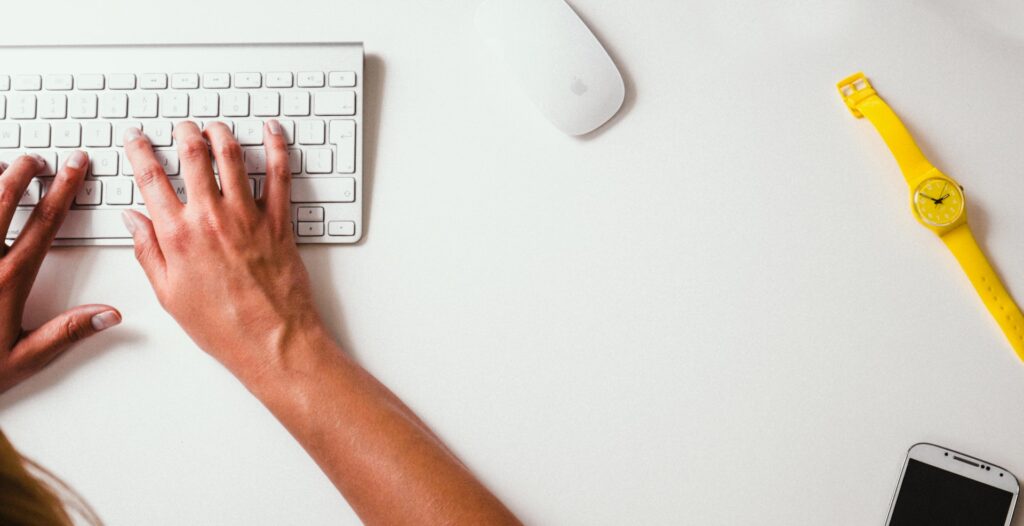 A women's hand tryping on a keyboard next to a mouse.