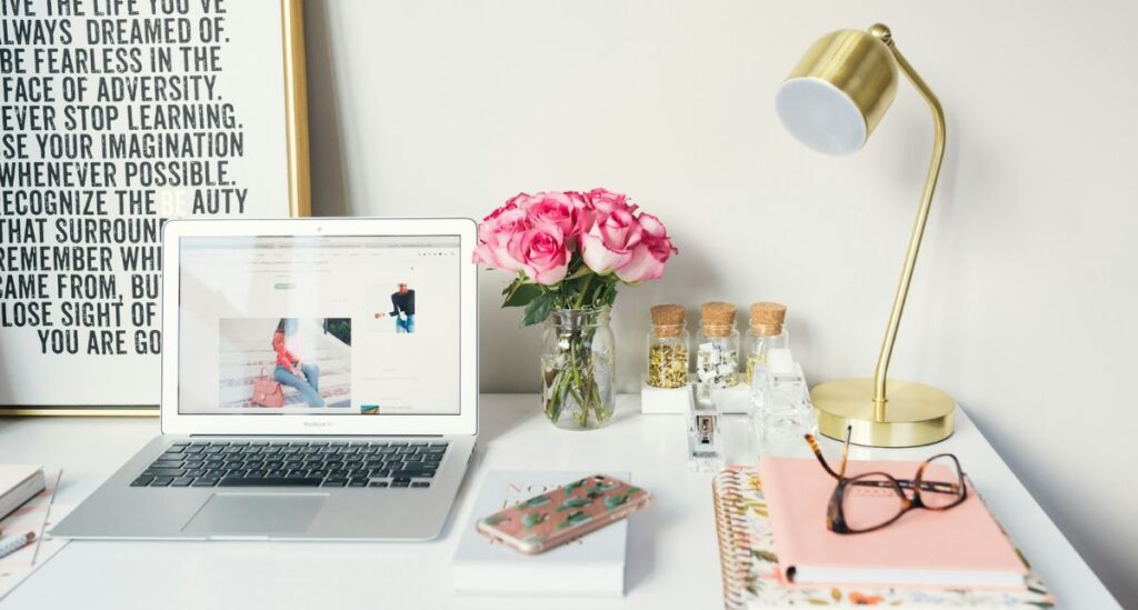 Many things on a white desk: flowers, a laptop, notebooks, a phone, and glasses.