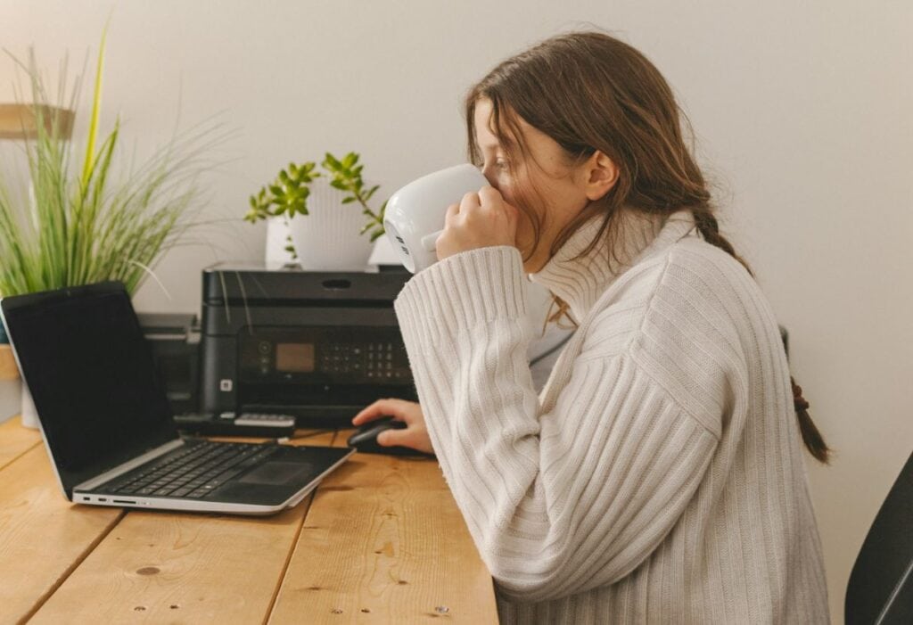 A woman sitting at a desk with a cup to her mouth while she works at a computer. 